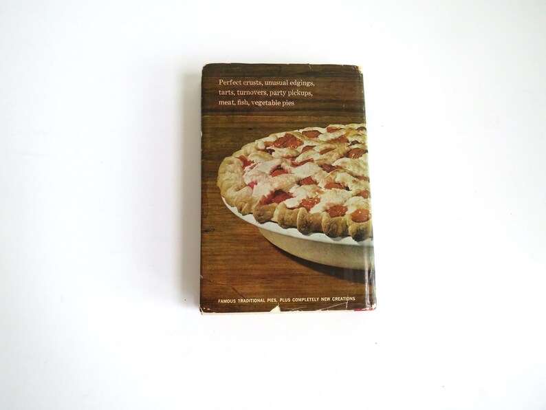 Puede incluir: Una portada de libro de cocina vintage con un primer plano de una tarta de cerezas con una corteza escamosa. El texto en la portada dice "Perfect crusts, unusual edgings, tarts, turnovers, party pickups, meat, fish, vegetable pies. Famous traditional pies plus completely new creations."