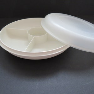 Puede incluir: Una bandeja de servicio de plástico blanco con tapa. La bandeja está dividida en cinco secciones, con un compartimento central circular y cuatro segmentos en forma de tarta. La tapa está parcialmente abierta.