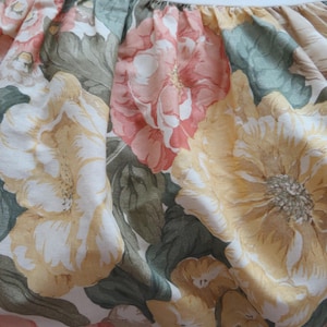 May include: A close-up of a floral fabric with a white background and large yellow, pink, and green flowers. The fabric has a ruffled edge.