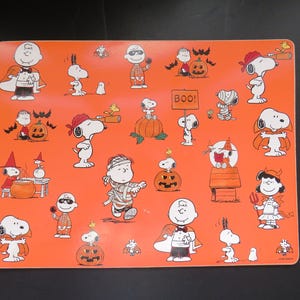 May include: Orange placemat featuring Halloween-themed Peanuts characters. Snoopy, Charlie Brown, and other characters are dressed in costumes, with pumpkins and bats. The word "BOO!" is on a sign.
