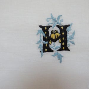 May include: White linen fabric with an embroidered letter "H". The letter is black with yellow accents and surrounded by light blue floral embroidery. The fabric has a decorative stitched edge.