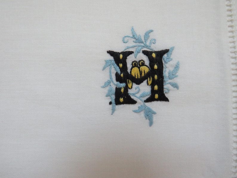 May include: White linen fabric with an embroidered letter "H". The letter is black with yellow accents and surrounded by light blue floral embroidery. The fabric has a decorative stitched edge.