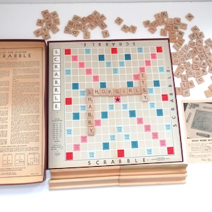 May include: A vintage Scrabble board game set, featuring a game board with the word "SHOP, GIRLS" spelled out. Includes wooden letter tiles, a box with rules, and a small card. The board has red, blue, and pink squares.