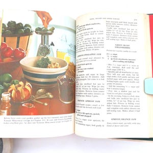 May include: An open vintage cookbook with a page featuring recipes for jams, jellies, and preserves. The left page displays a colorful illustration of a food grinder, a mixing bowl, and fresh produce. The right page contains printed text recipes.