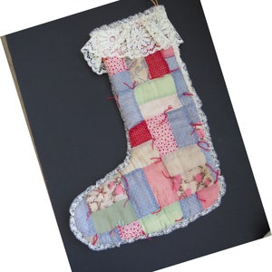 Può includere: Una calza di Natale fatta a mano con un design patchwork. La calza presenta quadrati di vari tessuti nei toni del blu, rosso, rosa e crema, con un bordo in pizzo bianco. Nastri rossi accentuano il patchwork e un pizzo color crema rifinisce la calza.