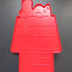 May include: A red plastic cookie cutter in the shape of Snoopy's doghouse, with Snoopy lying on top. The cutter has a rectangular base with horizontal lines and a doghouse roof. The design is a classic cartoon character.