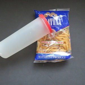 May include: A clear plastic container with a red lid is on top of a blue and white bag of pasta. The bag is labeled "ATINA" and "PENNE RIGATE NO. 86". The pasta is a light brown color.