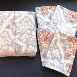 May include: A folded set of light blue floral patterned fabric, next to a set of unfolded fabric with the same design. The pattern includes white flowers and accents of orange and brown. The fabric is likely for home decor.