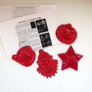 Puede incluir: Cortadores de galletas rojos vintage con forma de estrella, calabaza, Papá Noel y payaso. Los cortadores están junto a una hoja de papel impresa con el título "Aunt Chick's 3-D Cookie Cutters".