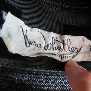 May include: A white label sewn onto a black hat with the text "Vera Whistler Original" in black cursive writing.