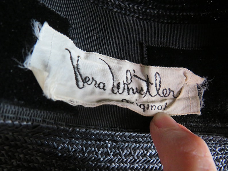 May include: A white label sewn onto a black hat with the text "Vera Whistler Original" in black cursive writing.