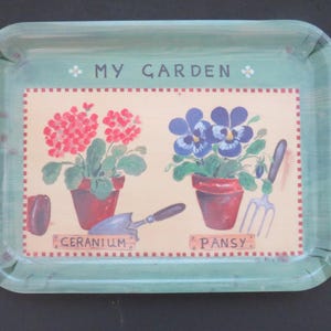 May include: A rectangular light green tray featuring the text MY GARDEN. The center illustration shows a red geranium in a pot with a trowel and a blue pansy in a pot with a hand fork, labeled GERANIUM and PANSY on a cream background.