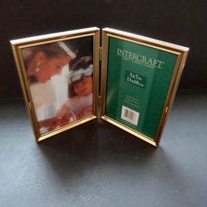 May include: A gold-colored, hinged picture frame with a beaded edge. One side displays a photograph of a woman and child, while the other side features a green insert with the text "INTERCRAFT" and dimensions of 5x7 inches (13x18cm).