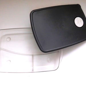 May include: A clear plastic food container with a black lid. The lid has a white button on the top.