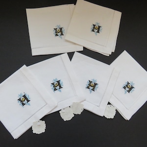 May include: A set of white linen napkins, each embroidered with a floral design around the letter "H". The embroidery features blue, yellow, and black accents. The napkins are neatly folded and arranged on a dark surface.