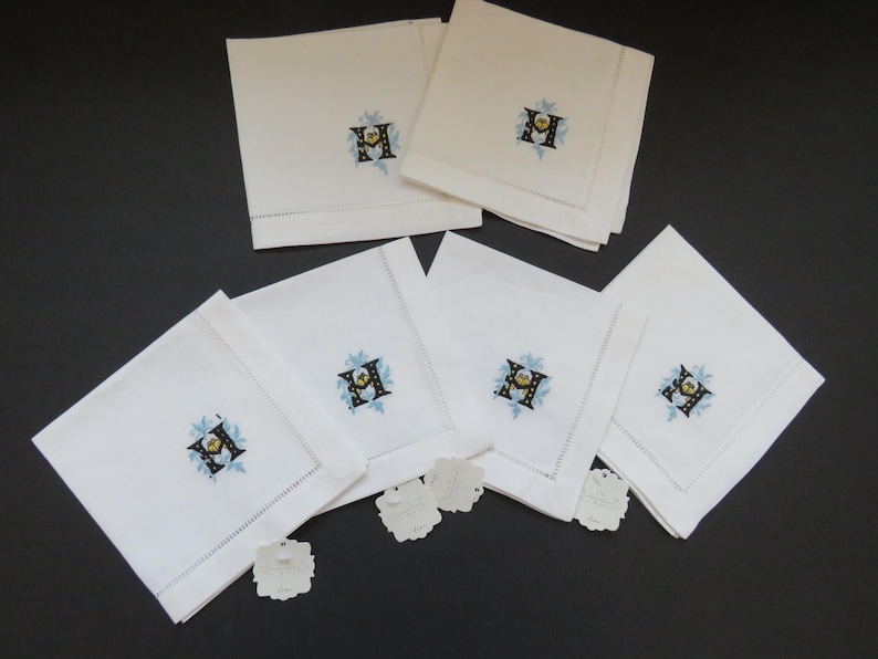 May include: A set of white linen napkins, each embroidered with a floral design around the letter "H". The embroidery features blue, yellow, and black accents. The napkins are neatly folded and arranged on a dark surface.