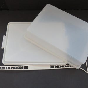 May include: A rectangular white plastic butter dish with a semi-translucent lid and a separate white plastic spreader tool featuring a long handle with circular cutouts and a flat, wide blade end, designed for serving and storing butter.