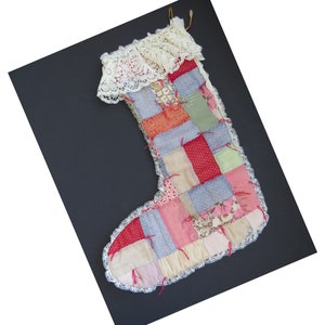 Può includere: Una calza di Natale patchwork con finiture e parte superiore in pizzo color crema. La calza è realizzata con vari quadrati di tessuto rossi, rosa, blu e verdi, con finiture in pizzo bianco. Un cordino dorato è attaccato per appenderla.