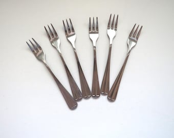 May include: Six silver forks with a decorative pattern on the handle. The forks are arranged in a fan shape on a white surface.