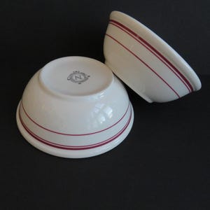 May include: Two white ceramic bowls with a maroon stripe design. One bowl is upright, displaying the bottom with a logo, while the other is tilted. The bowls are likely for serving or mixing.