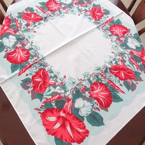 May include: White tablecloth with a vibrant floral design featuring large red and white striped flowers, green leaves, and a light blue border. The tablecloth is square and folded on a round wooden table.