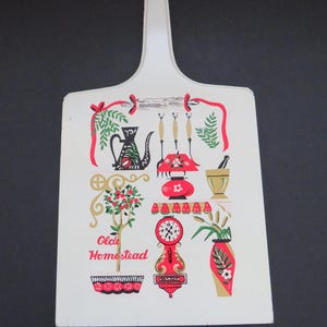 May include: A white cutting board with a handle, featuring a vintage kitchen scene. The design includes a red and black clock, a vase with flowers, and the words "Olde Homestead" in gold lettering. Red ribbons and green foliage complete the retro aesthetic.