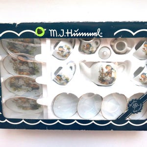 Op de afbeelding: Een vintage M.J. Hummel porseleinen theeset in de originele doos. De set bevat een theepot, melkkan, suikerpot, kopjes en schotels, allemaal versierd met Hummel-beeldjes. De doos is marineblauw met een witte geschulpte rand.
