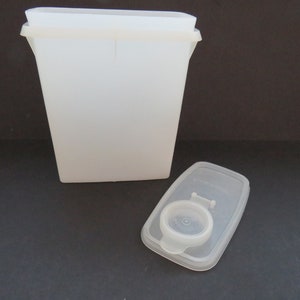 Puede incluir: Un recipiente de plástico blanco con tapa. El recipiente tiene forma rectangular y un asa en la parte superior. La tapa también es blanca y tiene una pequeña asa en la parte superior.