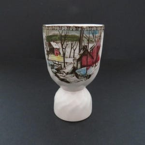 May include: A white ceramic egg cup with a detailed winter landscape scene. The cup features a flared top with a gold rim and a pedestal base with a spiral design. The artwork depicts red buildings, trees, and snow.
