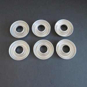 Pode incluir: Seis pequenas formas redondas de alumínio para bolo bundt, dispostas sobre uma superfície cinza escuro. Cada forma tem um orifício central e um design de anéis concêntricos, ideal para assar.