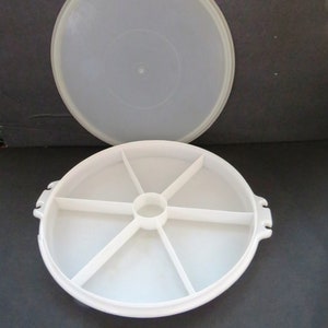 Puede incluir: Bandeja de servir blanca de plástico dividida con tapa. La bandeja tiene seis secciones y un círculo central. La tapa es transparente.