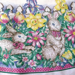 May include: A white tablecloth with a pink and white polka dot border. The tablecloth features a floral design with bunnies, tulips, daffodils, and other flowers in shades of pink, yellow, blue, and green.