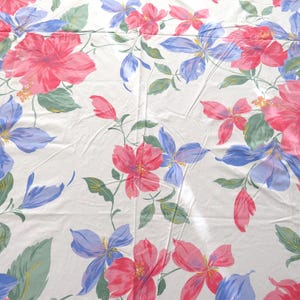 May include: White tablecloth with a floral pattern featuring large pink and red hibiscus flowers, blue iris-like flowers, and green leaves. The flowers are scattered across the fabric, creating a vibrant and colorful design.