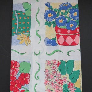 May include: A white tablecloth with a colorful floral pattern. The design includes red, pink, and blue flowers in pots and scattered across the fabric. Green leaves and stems are also present, creating a vibrant, cheerful aesthetic.