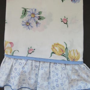 Puede incluir: Sábana blanca con estampado floral con flores azules, amarillas y rosas. La sábana tiene un ribete de volantes azules.