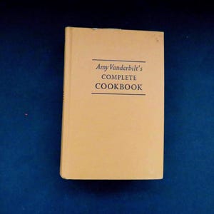 May include: A vintage cookbook titled "Amy Vanderbilt's Complete Cookbook" with a light tan cover. The title is printed in black, centered on the cover. The book is set against a dark blue background.
