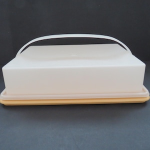 以下が含まれることがあります： オフホワイトの長方形のプラスチック製バターディッシュ。蓋とベースは淡い黄色で、白いアーチ型のハンドルが付いています。標準サイズのバターを収納できるように設計されています。