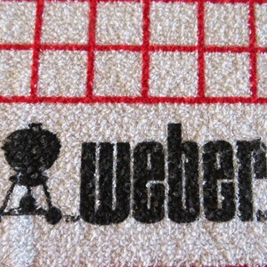 May include: White kitchen towel with a red grid pattern. The towel has a black silhouette of a grill and the word "Weber" printed on it.