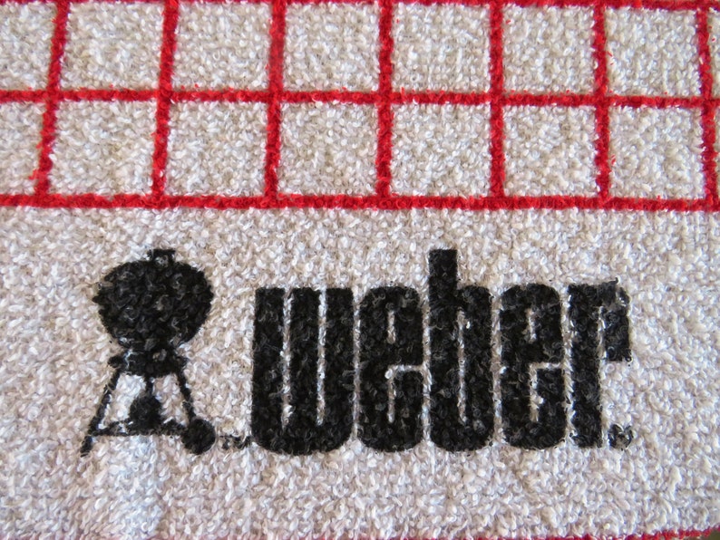 May include: White kitchen towel with a red grid pattern. The towel has a black silhouette of a grill and the word "Weber" printed on it.
