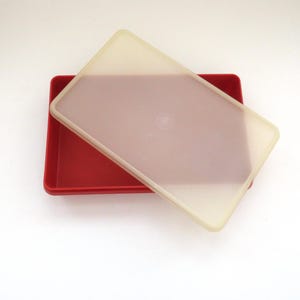 Puede incluir: Un recipiente rectangular rojo con una tapa translúcida blanquecina. La tapa está ligeramente entreabierta, revelando el interior del recipiente. El diseño es simple y funcional.