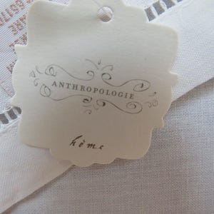 May include: A cream-colored tag with the word "ANTHROPOLOGIE" in a decorative font and the word "home" in script. The tag is scalloped around the edges and is attached to a white fabric with a lace trim.