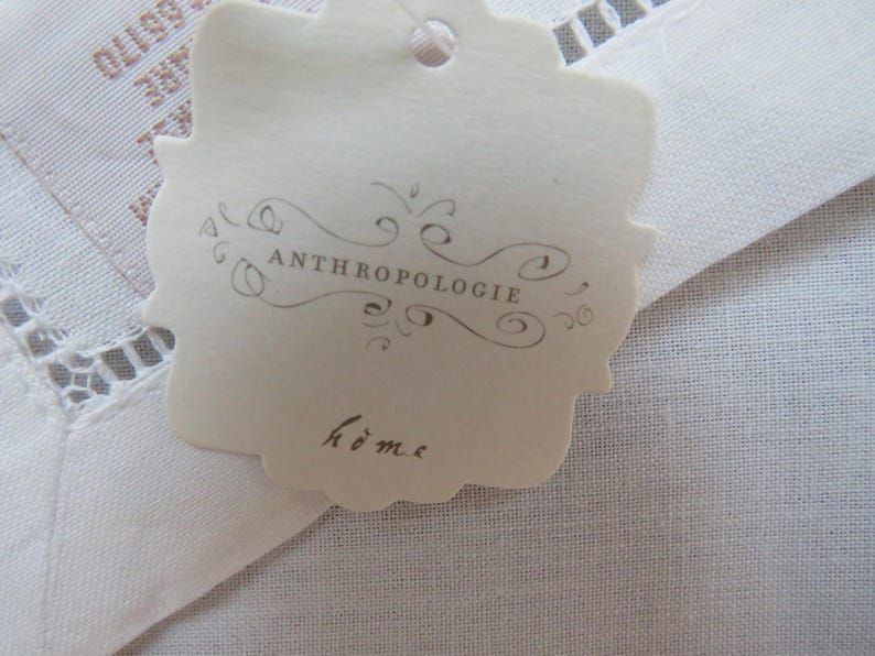 May include: A cream-colored tag with the word "ANTHROPOLOGIE" in a decorative font and the word "home" in script. The tag is scalloped around the edges and is attached to a white fabric with a lace trim.