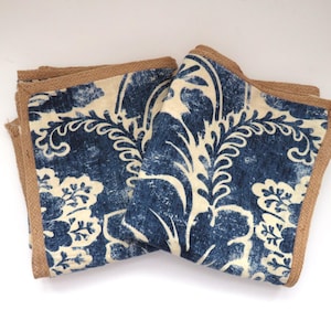 Op de afbeelding: Twee gevouwen vierkante servetten met een blauw en crèmekleurig bloemmotief. De servetten hebben een jute rand. Het bloemmotief is een gestileerd damastpatroon. De servetten zijn op elkaar gestapeld.