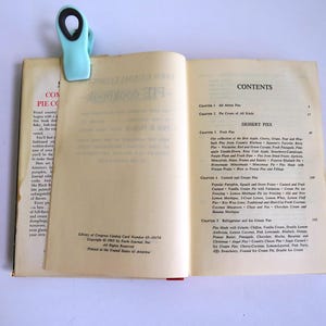 Puede incluir: Un libro abierto con un clip de carpeta azul en la p&aacute;gina superior. El libro se titula "Complete Pie Cookbook" y la p&aacute;gina de contenido enumera diferentes tipos de tartas, incluidas tartas de frutas, tartas de crema y tartas de refrigerador.