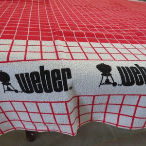 May include: A red and white checkered kitchen towel with the Weber logo and a silhouette of a grill. The text "Weber" is printed in black.
