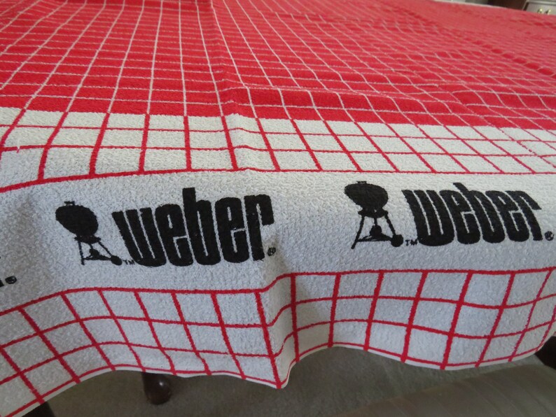 May include: A red and white checkered kitchen towel with the Weber logo and a silhouette of a grill. The text "Weber" is printed in black.