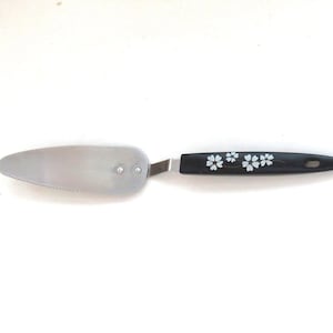 May include: A cake server with a metallic silver triangular blade featuring a serrated edge and two rivets. The handle is black with a white floral pattern consisting of three small flowers and a hanging hole at the end of the handle.