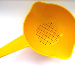 May include: A bright yellow plastic colander with a long handle featuring a circular hole at the end. The bowl of the colander has a flat bottom with a cluster of small, evenly spaced drainage holes in the center for straining liquids.