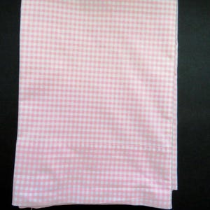 May include: A rectangular fabric piece featuring a light pink and white gingham check pattern. The textile is folded neatly, displaying a consistent grid design across the entire surface with a straight hem visible along the bottom edge.