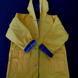 Könnte beinhalten: Ein leuchtend gelber Regenmantel mit Kapuze und Taschen. Die Manschetten haben ein blau-rotes Karomuster. Das Innenfutter ist blau. Der Mantel hat eine Knopfleiste.